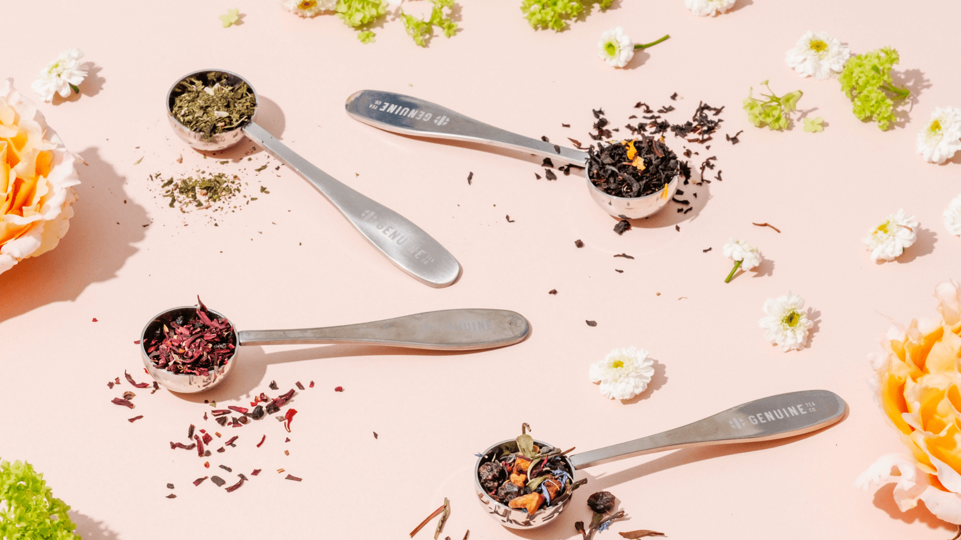 Four metal teaspoons labeled “Genuine Tea Co.” filled with different loose leaf teas on a soft pink background, surrounded by small white flowers and an orange rose.